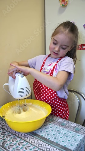 Adorable young girl in a red polka-dot apron happily operates a white electric hand mixer in a large yellow bowl with pale yellow batter. Focused and joyful moment of a child helping in the kitchen. 