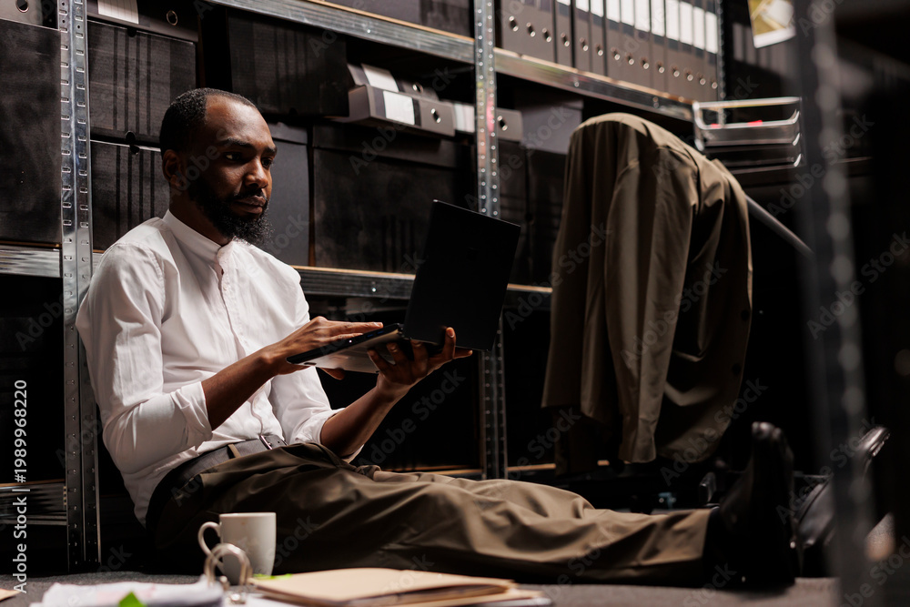 Naklejka premium Private detective conducting investigation using laptop while sitting on floor surrounded by police report papers. Detective analyzing crime case information on computer in evening
