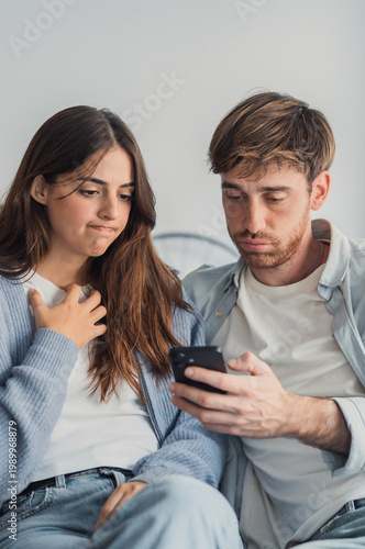 Foto A young couple sits closely on a couch, both looking worried and upset as they view messages on a smartphone, conveying distrust, jealousy and strained communication in a domestic setting