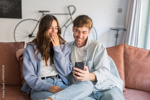 Schilderij op canvas Young couple sits close on a sofa in a cozy living room, smiling and laughing as they look at a smartphone together, sharing a light, intimate and joyful moment at home