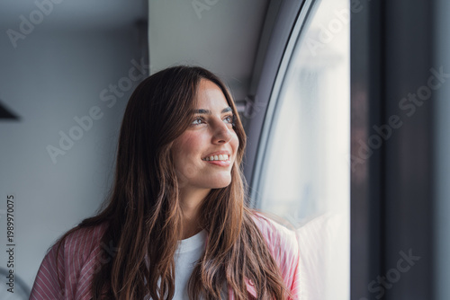 Obraz na plátne Young woman with long hair smiles as she gazes out a window, bathed in soft natural light