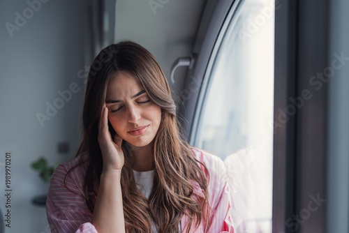 Tableau sur toile Young woman sits by a window with her hand on her temple, showing headache, stress and fatigue in a quiet, introspective moment at home