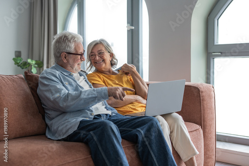 A happy senior couple sits on their living room sofa sharing a laptop, smiling and pointing as they browse, video call or manage online tasks together in a warm, comfortable home setting.