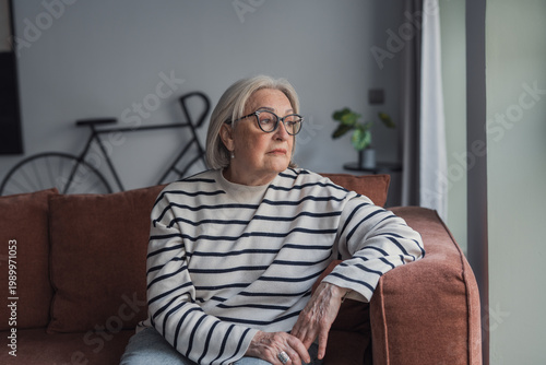 Fotografi A thoughtful senior woman with glasses sits on a sofa by a window at home, gazing outside with a reflective, calm expression in a cozy living room setting