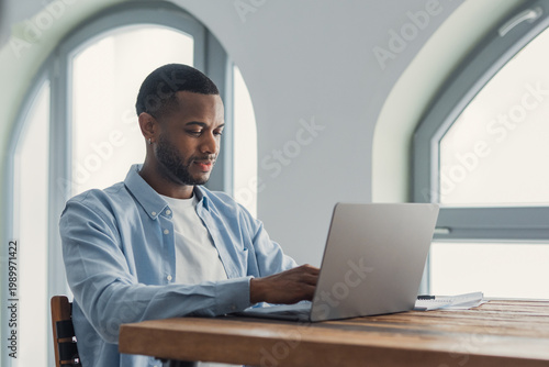 Young man working on laptop at home office by arched window, remote work and concentration
