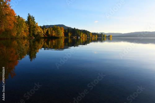 A tranquil image of a calm lake in late autumn with brightly colored trees lining the shoreline.  The trees are displaying an array of red, yellow, and orange colors.  