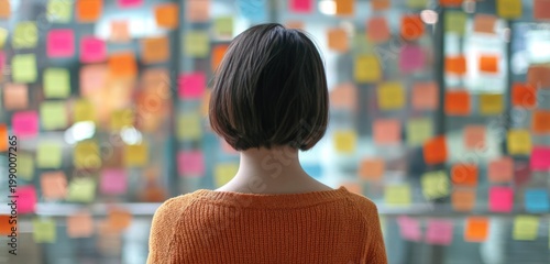 Asian woman with short hair wearing an orange sweater standing in front of a colorful wall covered with sticky notes and posters
