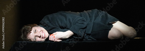 Fényképezés In this image, a woman lies on a dark stage, laughing joyously, embodying a carefree spirit, contrasting with earlier somberness and inviting positivity through her vibrant expression