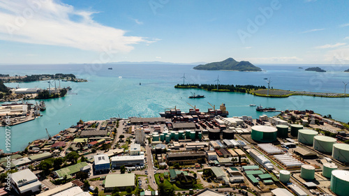 Harbor with industrial buildings, storage tanks, containers, and turquoise sea, with green islands in the background. Seychelles, Mahe. Victoria.