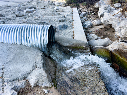 Water flows out of a corrugated metal pipe onto rocky concrete and stone surrounded by large boulders and sparse vegetation creating an energetic drainage scene outdoors