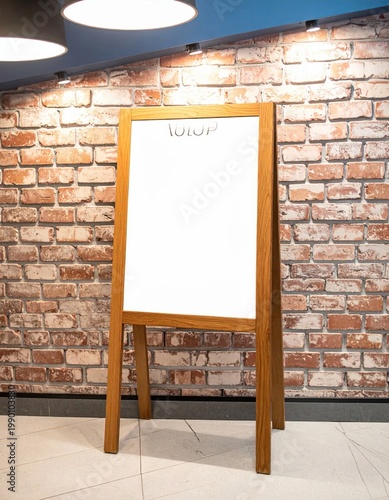 Wooden easel stand with a blank white board against a rustic brick wall, ideal for menu display, signage, advertising mockup or event announcements