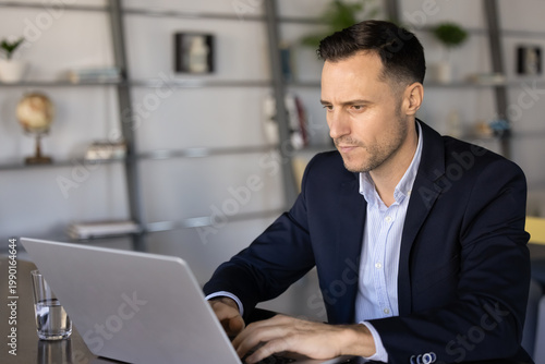 Focused businessman working on laptop sitting at office desk, responding on business email, reviewing digital documents or project information on screen, analyzing data, preparing report at workplace