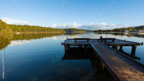 A boat and fishing jetty in perspective juts out into the calm waters of a lake with reflections of blue sky, clouds and trees in the water at Burrill Lake in New South Wales, Australia.