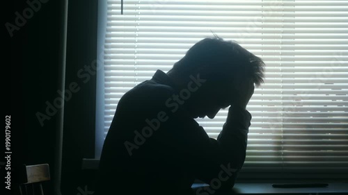 Sad and depressed man in silhouette sits alone at table in dark room, his head in his hands, representing despair, anxiety, loneliness, mental health issues. Depressed, sad man sitting in dark room