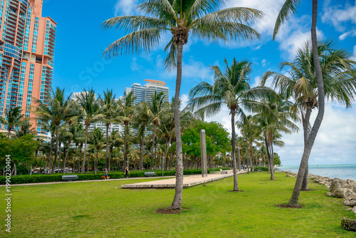 Miami, USA, 9 April 2026: Public park promenade with palm trees and residential towers. Wide view of a paved path in a tropical park with lush palm trees and tall city buildings under a blue sky.