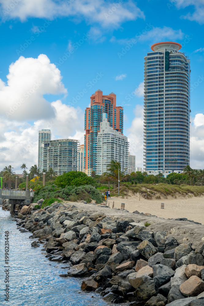 Fototapeta premium Vertical view of rock jetty and Miami skyscrapers. Large stones of a jetty leading towards luxury residential towers under a blue sky in Miami Beach.