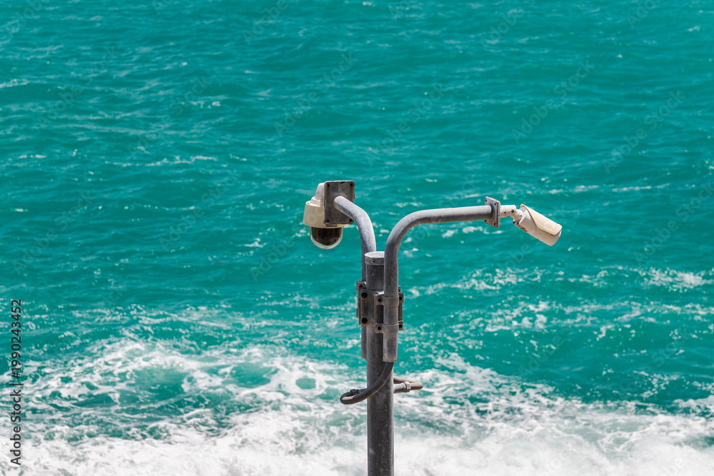 Fototapeta premium Security surveillance cameras on metal pole above turquoise sea with waves