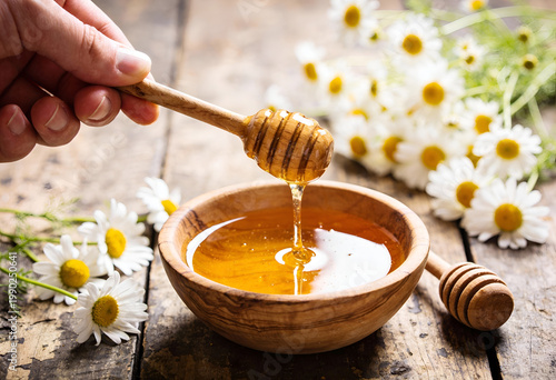 Hand drizzling honey from dipper into wooden bowl with flowers