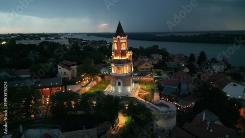 Dramatic drone orbit around illuminated Gardos tower at twilight with incoming lightning storm over Danube river. Storm clouds and lightning striking the background over Zemun, Belgrade, Serbia.