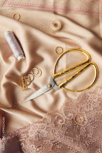 A close-up photo of tailor's tools: beige lace, spool, accessories for straps, decorations, scissors. Photo grain added.