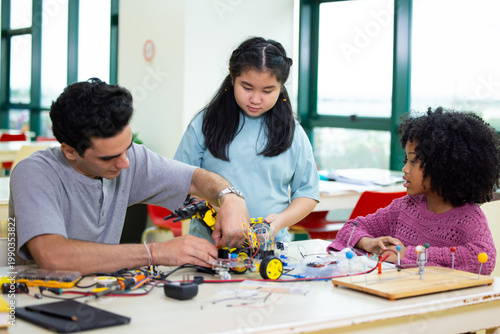 Young teacher guides students through hands-on robotic arm engineering project. Students focus intently, collaborating at shared workbench. Bright room, large windows. Education, Back to school