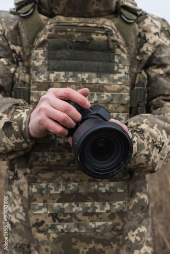 Military photographer in camouflage uniform holding professional camera outdoors. Documentary style image capturing field work, focus, and visual storytelling in real conditions.