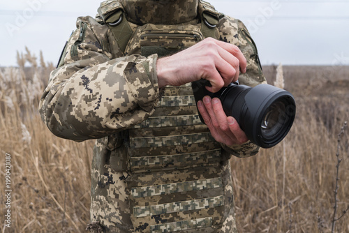 Military photographer in camouflage uniform holding professional camera outdoors. Documentary style image capturing field work, focus, and visual storytelling in real conditions.