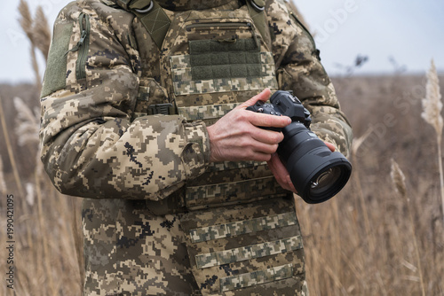 Military photographer in camouflage uniform holding professional camera outdoors. Documentary style image capturing field work, focus, and visual storytelling in real conditions.