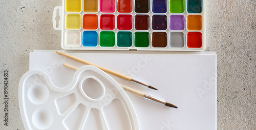 Colorful watercolor supplies are arranged on a table with brushes and blank paper ready for creativity in a bright space