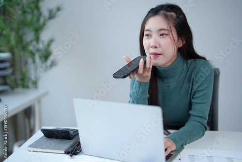 Schilderij op canvas Asian businesswoman multitasking at laptop and speaking on smartphone using voic