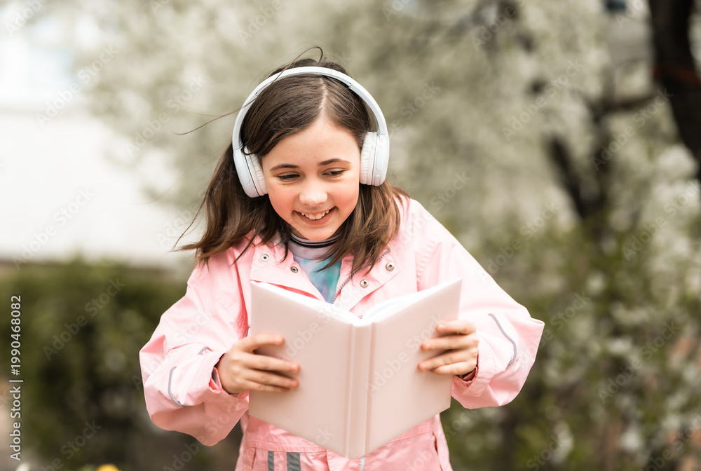 Fototapeta premium Happy 10-year-old girl using headphones to listen to an audiobook while reading in a blooming spring garden; concept of modern education and e-learning.
