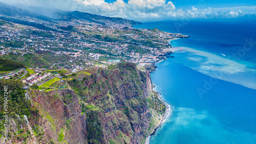 Aerial view of the dramatic cliffs of Cabo Girão, Madeira, with Funchal's urban sprawl stretching along the vibrant coastline under a clear sky. A stunning blend of nature and civilization.