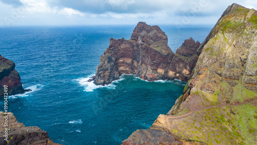 Rugged cliffs meet the Atlantic at Ponta de São Lourenço, Madeira. A dramatic coastal path winds along vibrant green and earthy rock formations under a cloudy sky.