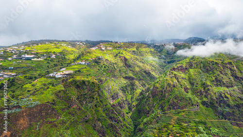 Dramatic aerial view of lush green terraced mountainsides dotted with charming villages, partially shrouded in atmospheric mist and clouds on the island of Madeira.Paul do Mar