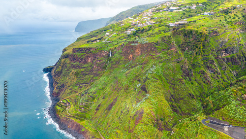 Aerial view of a picturesque coastal village and lush terraced cliffs on the vibrant island of Madeira, Portugal, under an overcast sky.