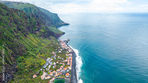 Aerial view of a picturesque Paul do Mar village clinging to Madeira's dramatic green cliffs, where the Atlantic waves meet the shore under a bright sky.