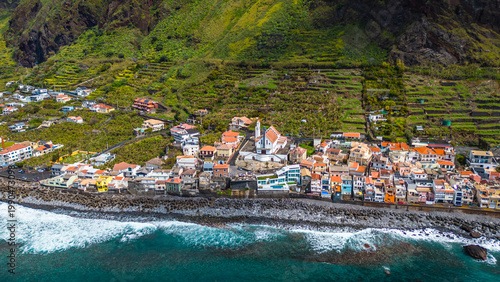 Aerial view of a vibrant Paul do Mar village nestled against lush green terraced mountains, with the Atlantic Ocean waves crashing on the rocky shore. Picturesque Madeira.