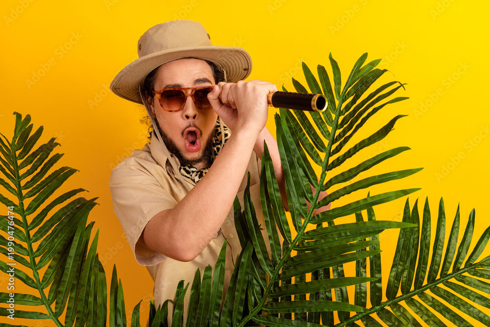 Naklejka premium Adventurous traveler with safari hat and mic among tropical leaves under a yellow backdrop
