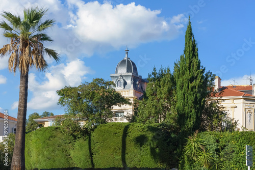 Cannes, France. Historic building with a dome surrounded by lush greenery and palm trees under a bright blue sky showcasing Mediterranean architecture
