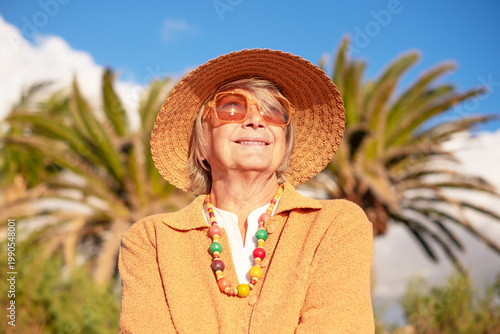 Portrait of smiling attractive senior woman in outdoors enjoying  a sunny day. Elderly lady in yellow and hat with palm trees on background