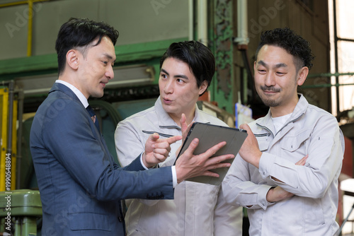 工場でタブレットを使い３人で打ち合わせする作業員とビジネスマンの現場、dxやIT、AI化の機械の導入や信金ビジネスマンが借り入れ提案のイメージ「それいいね！」