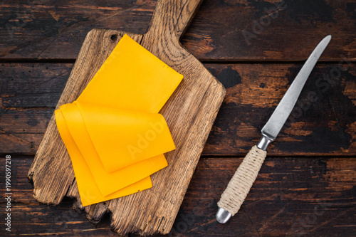 Sliced cheddar style cheese on wooden cutting board with knife over rustic table background and copy space for food design