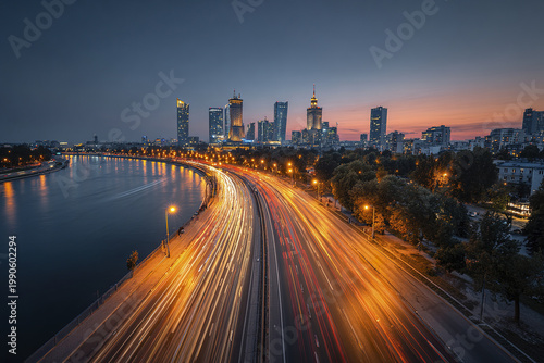 Panoramic city skyline at sunset with long exposure traffic light trails along riverfront highway, urban buildings, and glowing street lamps creating dynamic evening scene