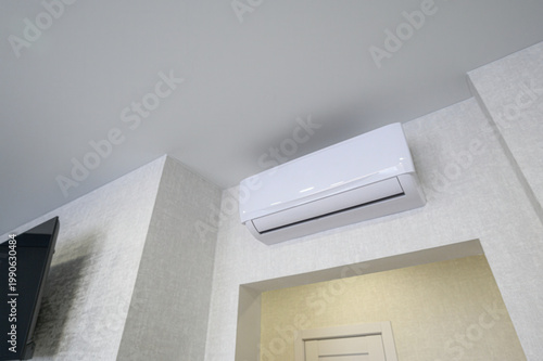 modern white split-system air conditioner mounted high on a light-textured wall, next to a TV and above a doorway in a minimalist interior