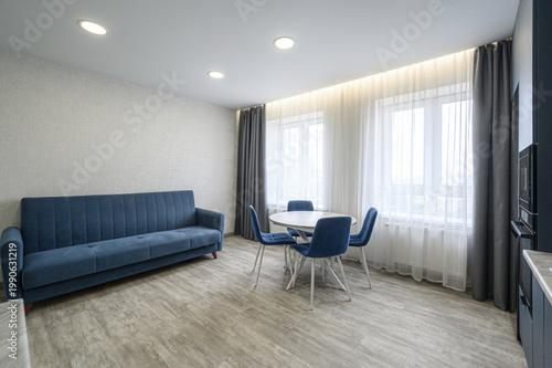 modern living room with a navy blue sofa, round dining table with four matching chairs, light wood flooring, sheer white curtains, recessed ceiling lights, and a kitchen area