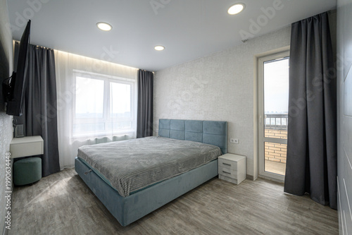 modern bedroom with a light blue upholstered bed, white bedside tables, grey curtains, large window, TV mounted on the wall, light grey flooring, and a balcony door