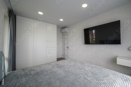 minimalist bedroom with a large white built-in wardrobe, wall-mounted TV, recessed ceiling lights, light grey textured walls, a neatly made bed with a grey quilt, and a small floating shelf.