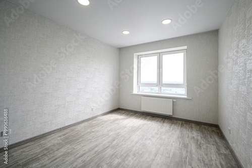 spacious, empty room with light grey textured walls, grey laminate flooring, a large double window with a white frame, a white radiator beneath it, and three recessed ceiling lights