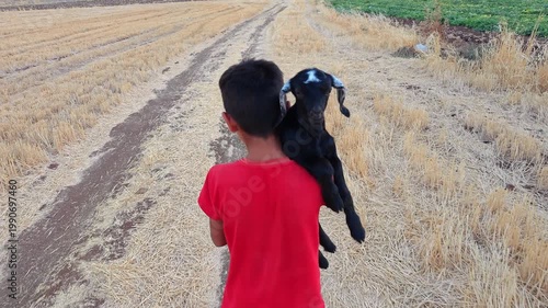 Close Up of Unrecognizable Boy Carrying Baby Goat on Shoulder on Rural Dirt Road
