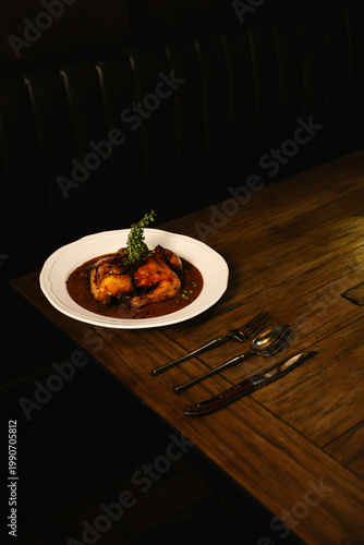Roasted chicken in rich sauce served on wooden table with cutlery in a warm, moody restaurant atmosphere.